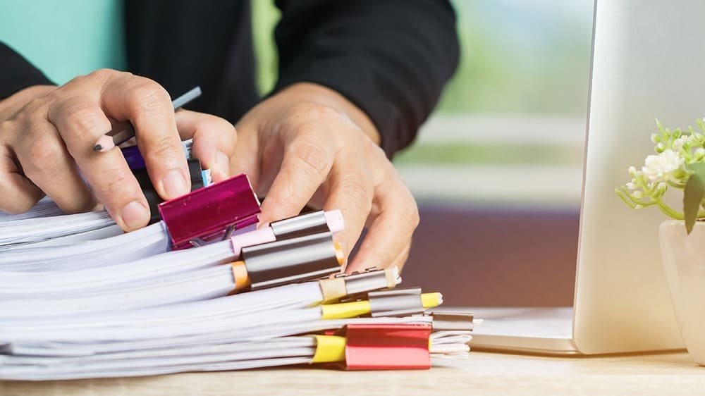 Person organizing documents with colorful binder clips on a desk next to a laptop and small plant