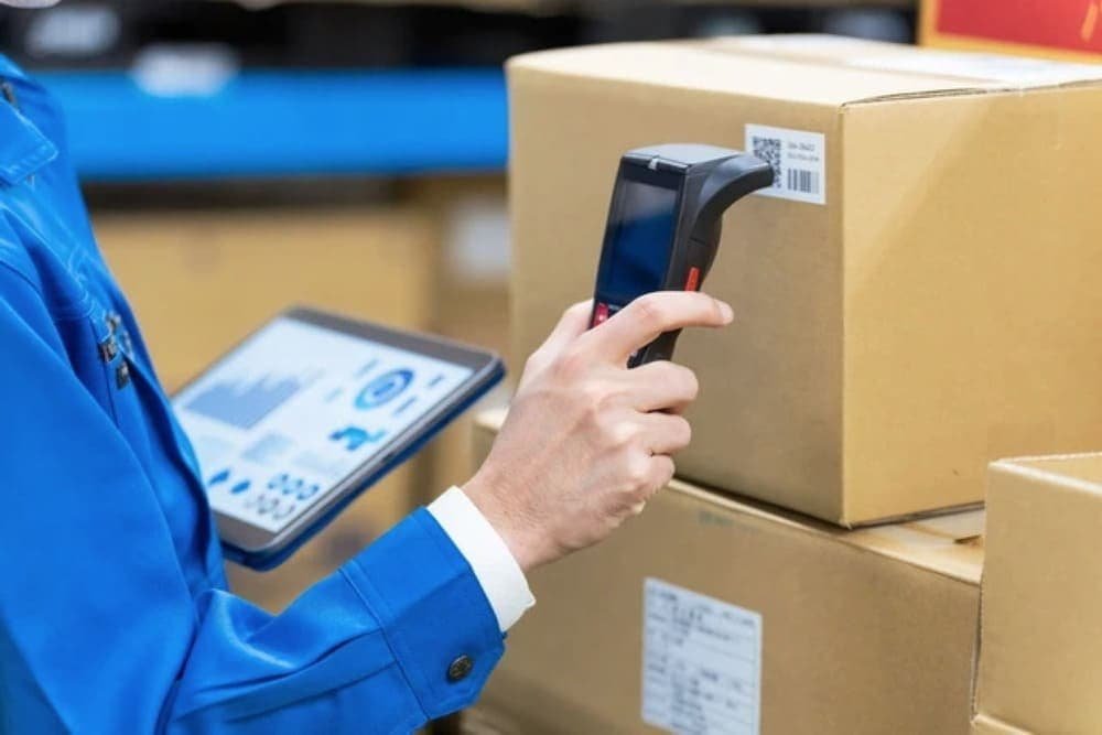 A person scans a barcode on a box with a handheld scanner, while holding a tablet displaying charts