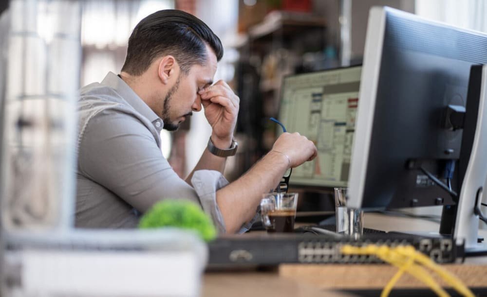 A man in an office rubs his eyes at a desk with computer monitors