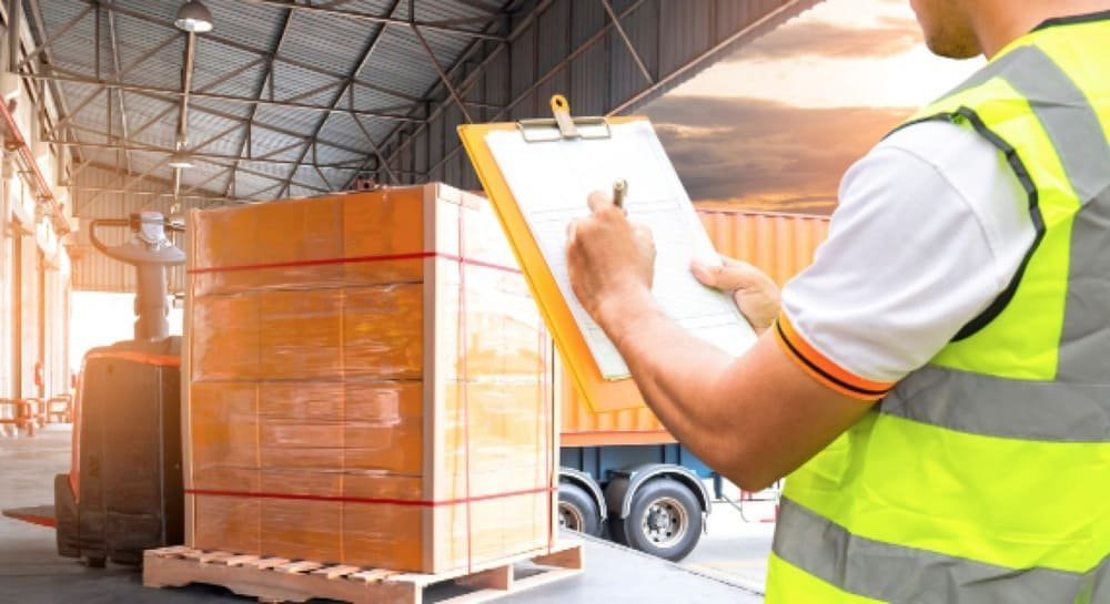 Worker with a clipboard inspecting a pallet of boxes on a forklift in a warehouse