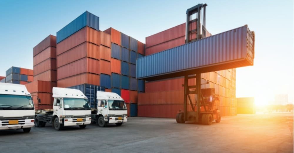 Shipping yard with stacked containers, semi-trucks, and a container handler lifting a blue container