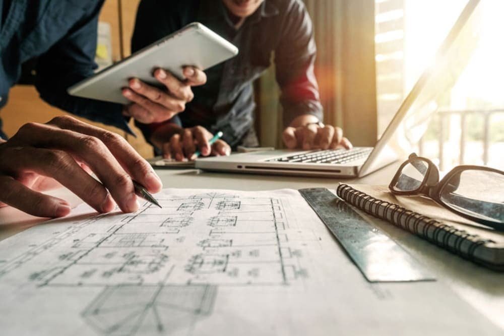 Two people working with a blueprint, laptop, and tablet in a sunlit workspace