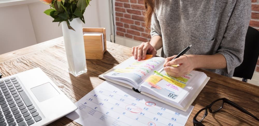 A person writing in a planner at a wooden table with a laptop, calendar, and flowers