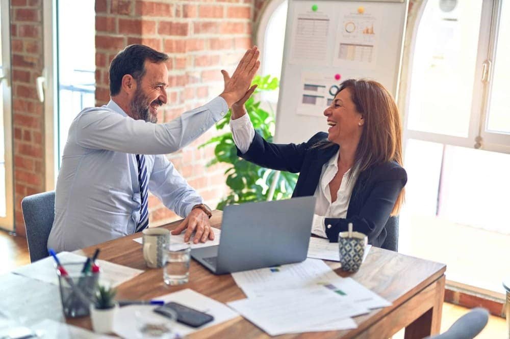 A man and woman high-fiving in a bright office with a laptop and documents on the table