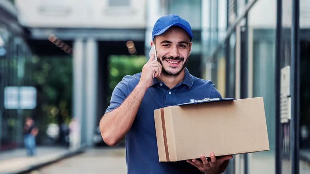 Man wearing a blue cap holding a cardboard box and talking on the phone in an urban setting