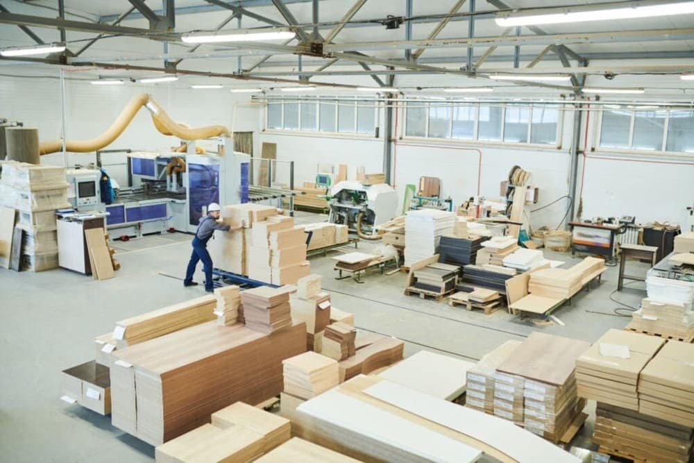 An industrial woodworking workshop with stacks of wooden panels, machinery, and a worker in a hard hat