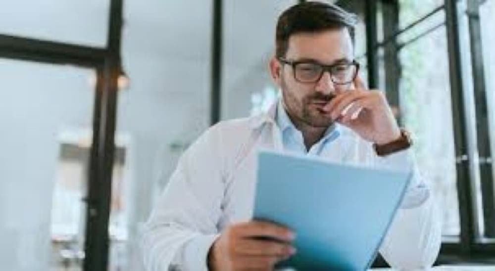 Man in glasses reviewing documents at a desk near bright windows
