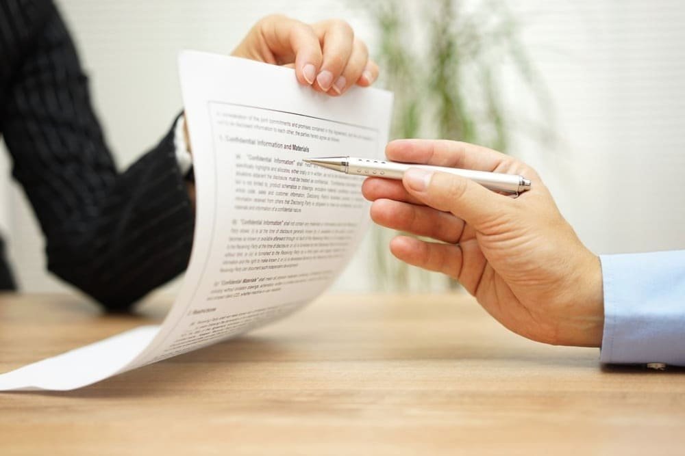 Two people exchanging a document and pen over a wooden desk for a business agreement
