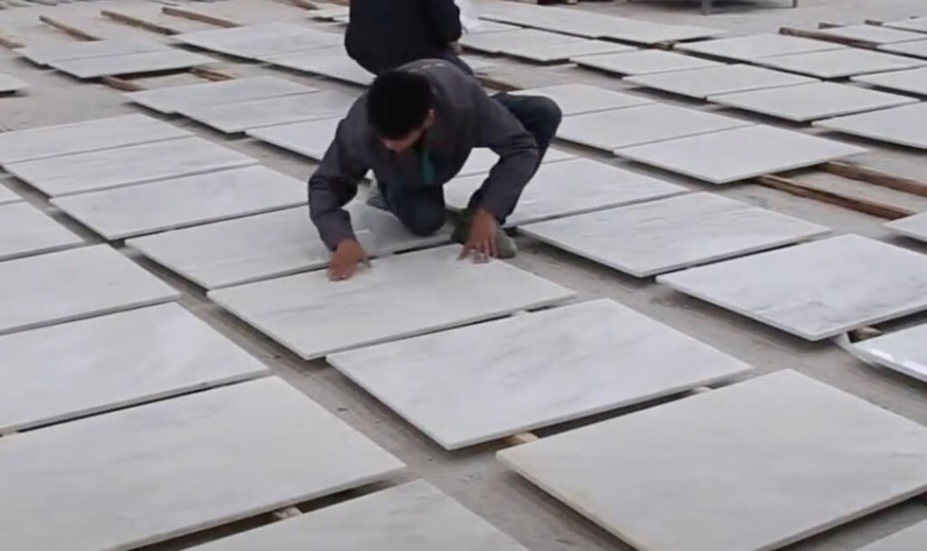 Person crouching outdoors arranging white marble floor tiles