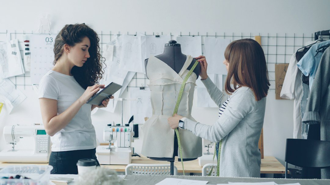 Apparel sample approval: garment makers measuring a garment on a dress form while recording specifications in a bright studio